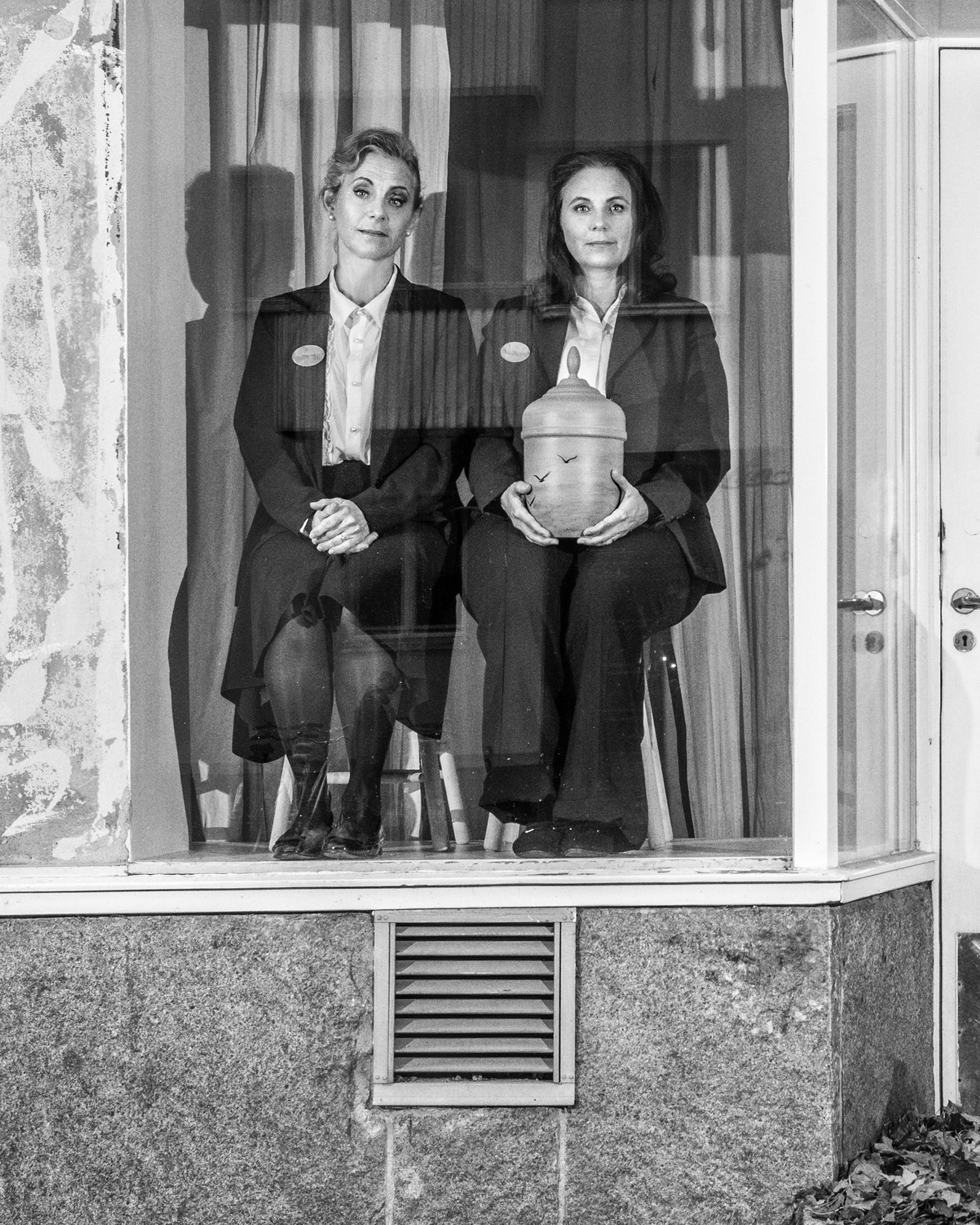 Helen Sjöholm och Anna Fahlstedt sitter på varsin pall i ett tomt skyltfönster. I sina händer har Anna en begravningsurna.