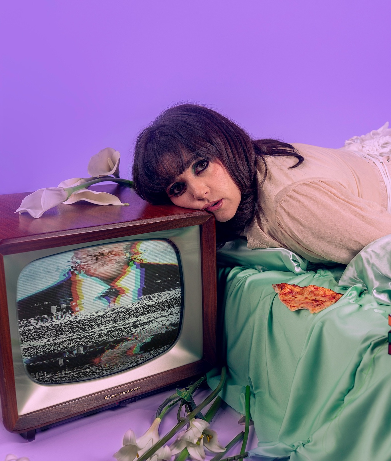 Kvinna ligger på säng med huvudet mot en tv med brus; pizza, Rubiks kub och blommor runtom i scen med lila bakgrund.