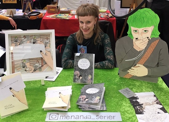 Serietecknaren Helena Menanda vid ett bord, med sina serier på. 