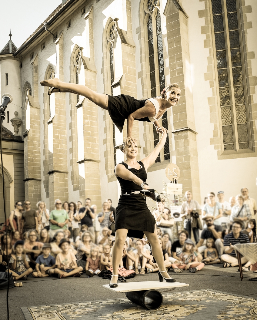 Två cirkusartister som utför akrobatik framför en kyrkbyggnad med publik