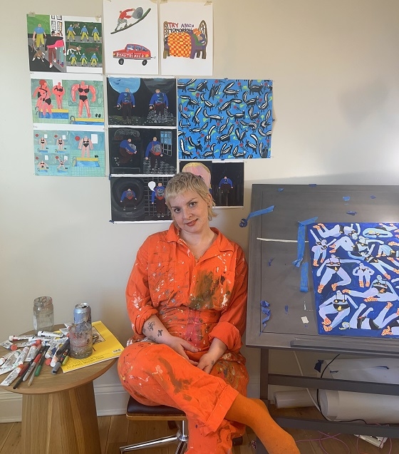 Serieskaparen Tara Booth sitter framför en vägg där flera av hennes verk är uppsatta. 