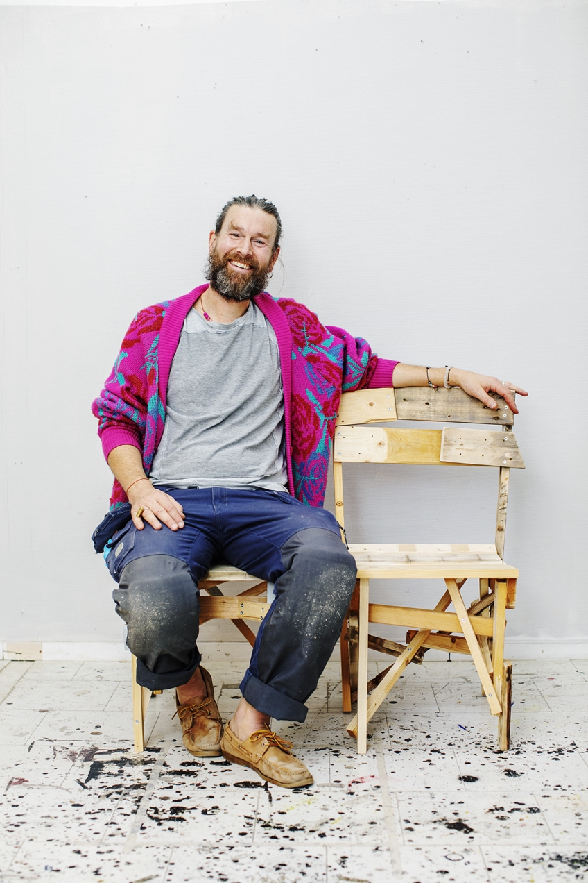 Möbelsnickaren Finn Ahlgren sitter på en möbel som han snickrat. Han ser glad ut och har en rosa skjorta och blå arbetsbyxor.