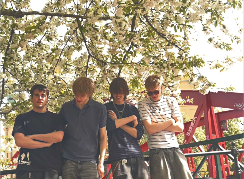 Fyra unga män i t-shirtar står under ett blommande träd framför en röd brokonstruktion utomhus.