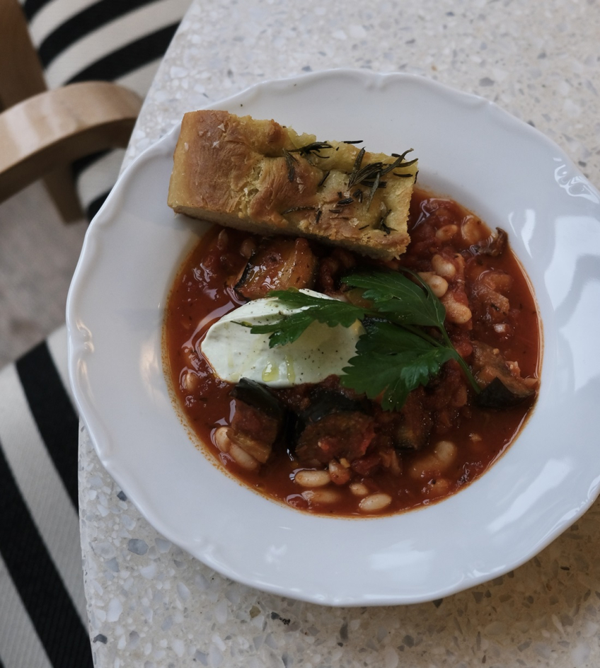 Tallrik med tomatbaserad gryta med vita bönor, zucchini, färskost, persilja och focacciabröd.