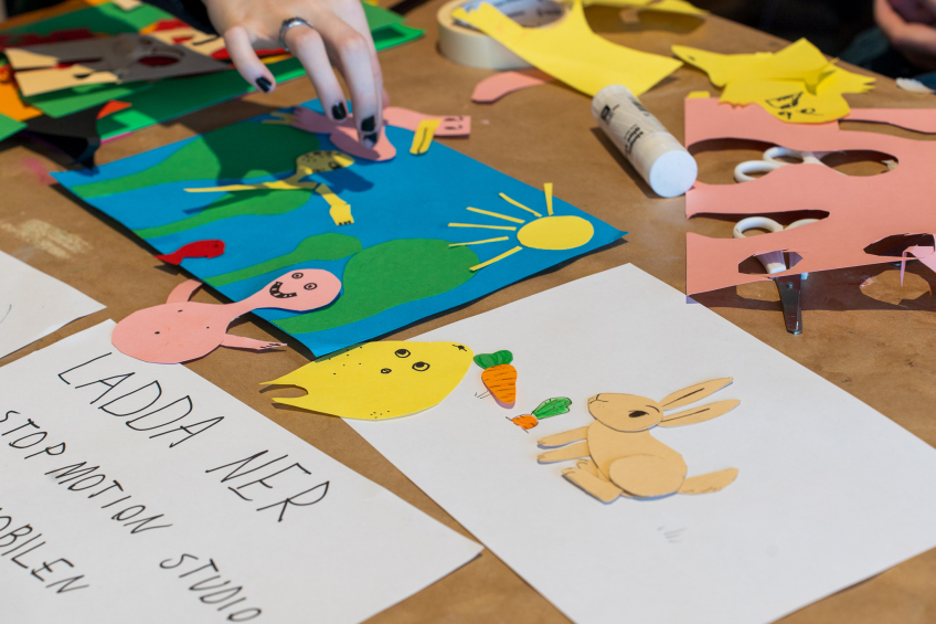 Hand skapar färgglada pappersfigurer för stop motion med sax, lim och utskurna djur på bordet.