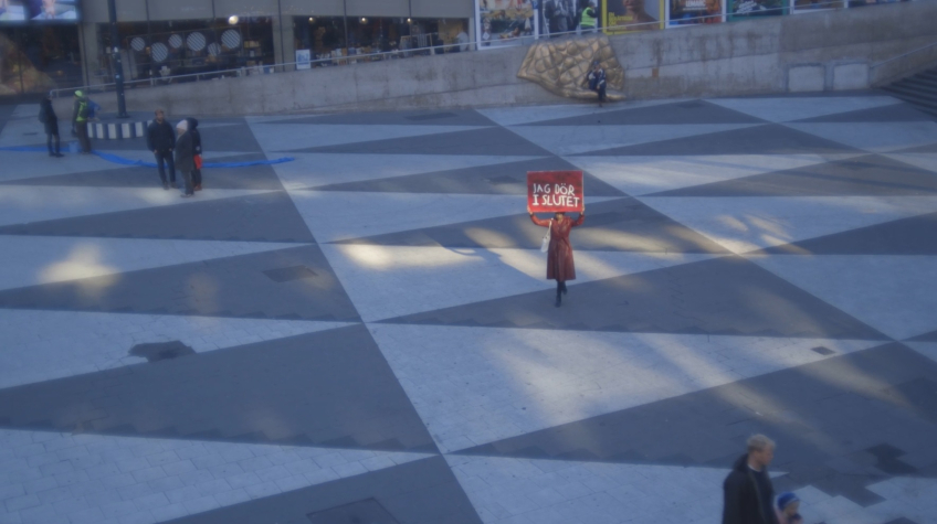 Person i röd kappa står ensam på torg och håller skylt med texten "JAG DÖR I SLUTET".