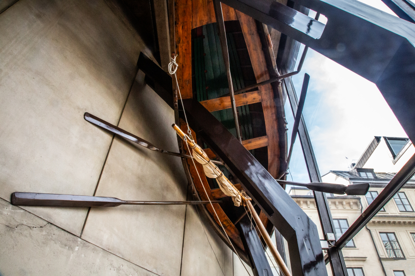 En stor träbåt sitter fastmonterad vertikalt på en byggnadsfasad, omgiven av mörka metallbalkar som bär upp konstruktionen. Åror och rep sträcker sig ut från båten, och genom glaspartierna syns himmel och intilliggande stadshus i bakgrunden.