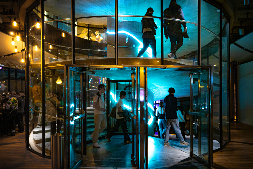 En grupp människor går in och ut genom en stor, glasad karusell­dörr i en modern byggnad på kvällen. Inne i entrén lyser starka turkosa och gula ljus som skapar reflexer i glaset. Några personer syns även i en spiraltrappa en våning upp.