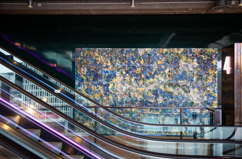 Ett stort konstverk i form av en mosaikliknande vägg bestående av blå, gröna, gula och vita strukturerade plattor sitter monterat vid rulltrapporna i Kulturhuset. Rulltrappornas glas och räcken skär diagonalt över bilden, och en ljusmarkering visar siffran 4 som anger våningsplanet. I bakgrunden syns utsikten mot stadens byggnader genom stora fönster.