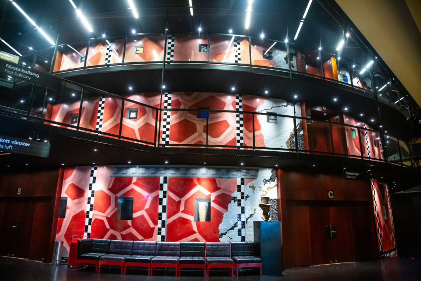 En stor teaterfoajé med två balkongplan syns i bild. Väggarna är täckta av röda och rosa geometriska mönster med svarta och vita rutband som vertikala accenter. Ljuspunkter i taket lyser upp de olika nivåerna, och längst ner i bild står en lång rad mörka sittbänkar. Dörrar märkta B och C leder vidare in i salongen.