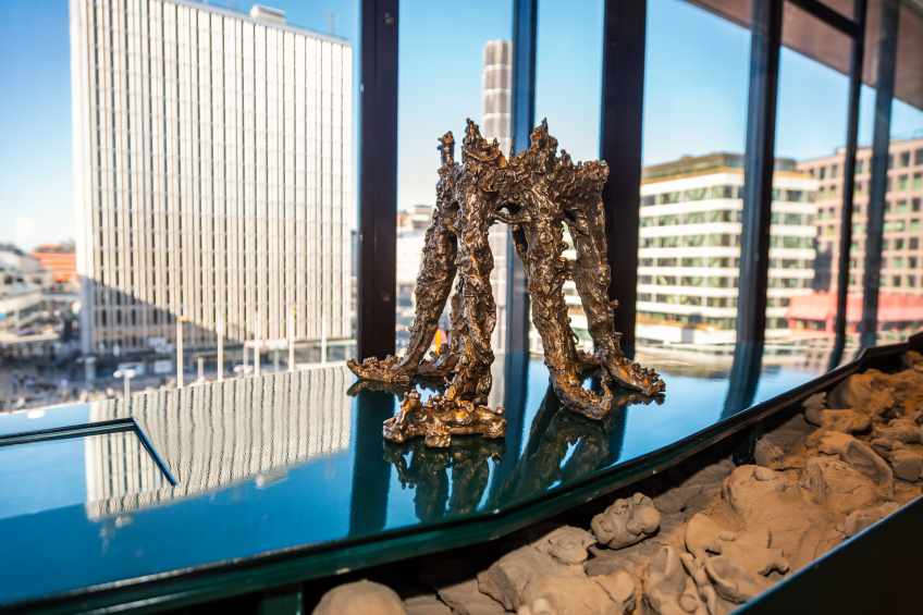 En liten bronsskulptur med organiska, grenliknande former står på en blank, mörkgrön yta framför stora fönster med utsikt över centrala Stockholm. Skulpturen består av flera knotiga pelarliknande ben som möts i ett gemensamt topparti. Nedanför står en installation av ljusbruna lerklumpar som fyller en lång behållare.