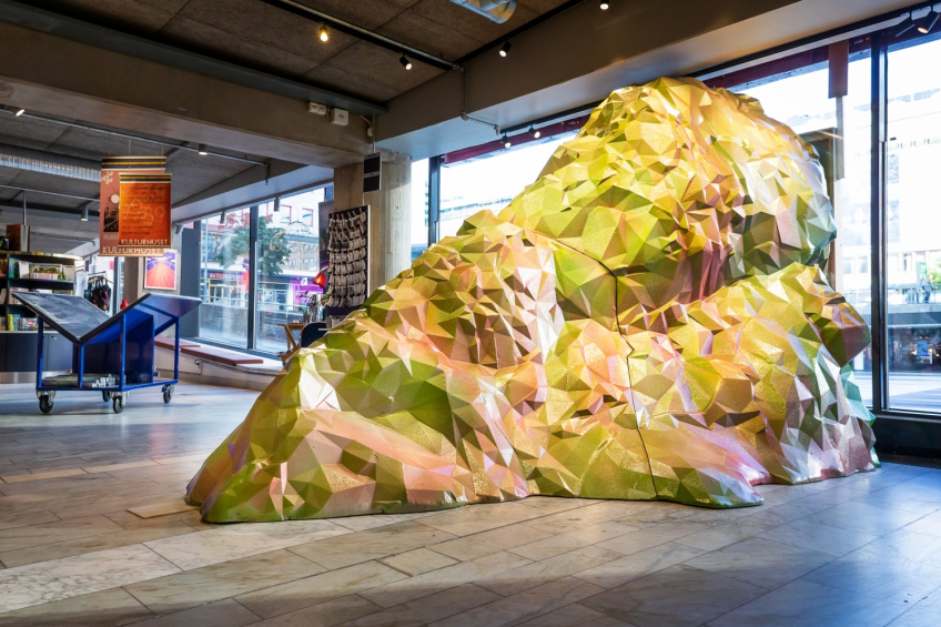 Ett stort, facetterat skulpturalt berg i skimrande gröna, rosa och gula toner står placerat i Kulturhusets ljusa entréplan. Verket har en geometriskt uppbyggd yta med många vinklar och reflektioner. I bakgrunden syns butiksmiljö, affischer och stora fönster mot stadsmiljön utanför.