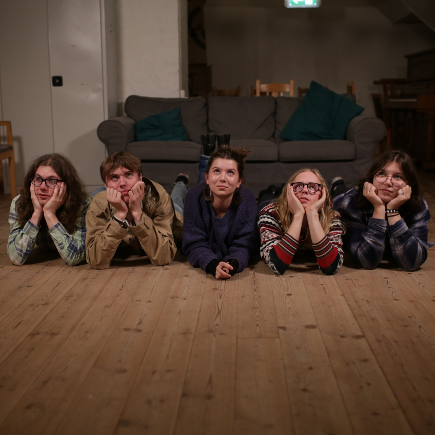 Fem unga personer ligger på golvet med hakan i händerna framför en soffa i ett vardagsrum.