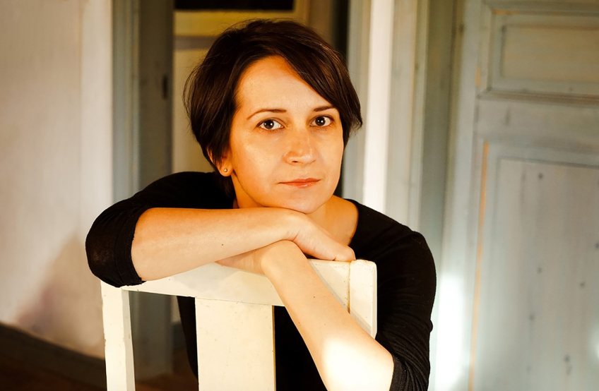 Kvinna ca 30–40 år med kort mörkt hår och svart långärmad tröja, lutar armarna på vit stol inomhus och ser mot kameran.