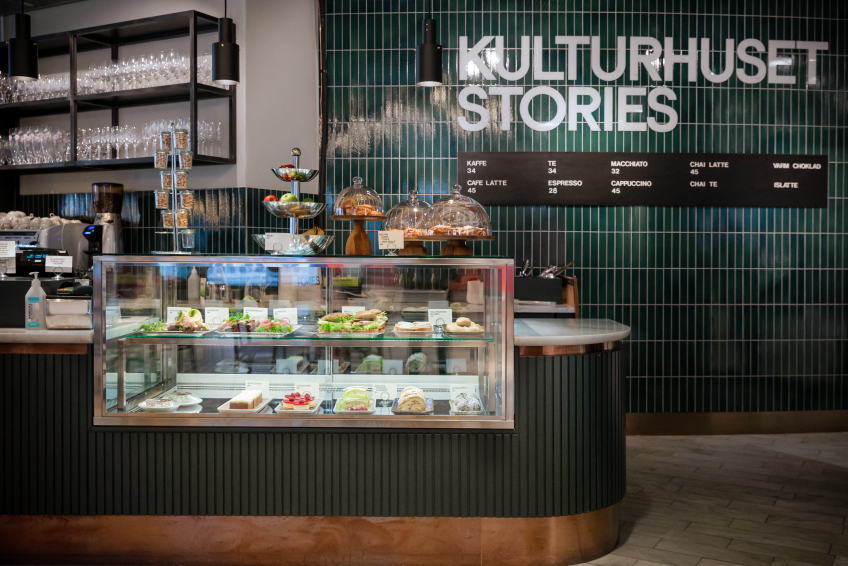 Cafédisk med smörgåsar och bakverk i glasmonter. Grön kakelvägg bakom med texten “Kulturhuset Stories” och meny.