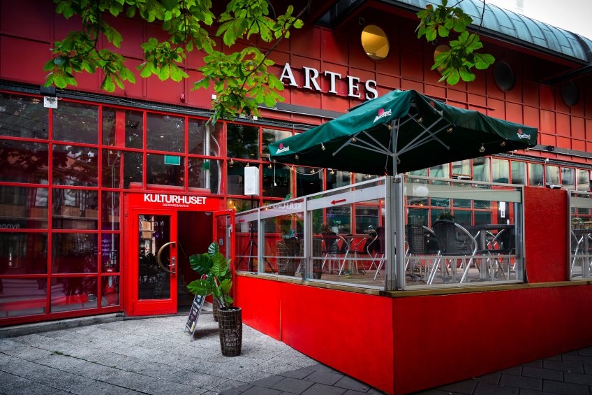 Uteservering utanför röd byggnad med skylten “Artes”. Bord och stolar under gröna parasoller vid entrén till Kulturhuset.