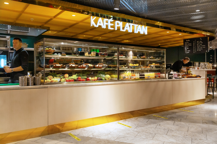Cafédisk med glasmontrar fyllda med mat och dryck. Skylt “Kaffe Plattan” ovanför, personal arbetar bakom disken.