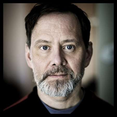 En medelålders man med skägg som tittar rakt in i kameran, med mjuk bakgrundsoskärpa.