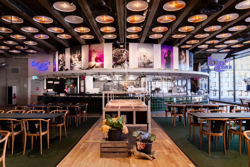 Restaurang med runda bord och stolar, öppen bar i mitten och fotokonst längs väggarna. Taket fyllt av runda lampor.