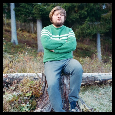 En person som sitter utomhus på en stubbe i skogsmiljö, med armarna i kors och ett mer allvarligt uttryck.