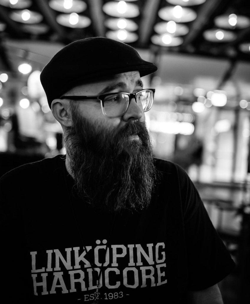 Svartvit porträttbild av en man med skägg, glasögon och keps, bär t-shirt med texten “Linköping Hardcore”.