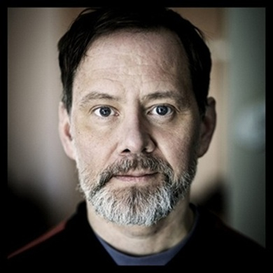 En medelålders man med skägg som tittar rakt in i kameran, med mjuk bakgrundsoskärpa.
