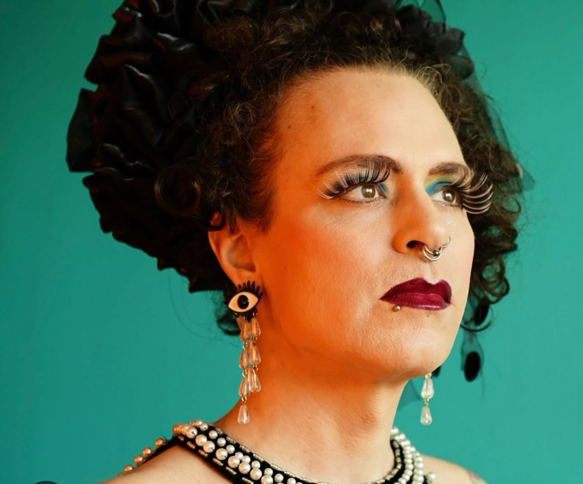 Porträtt av en person med dramatisk makeup, långa lösögonfransar och smycken. Personen blickar åt sidan mot en turkos bakgrund.