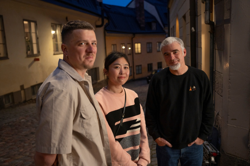 Tre personer står tillsammans på en kullerstensgård mellan hus i skymningsljus och tittar mot kameran.
