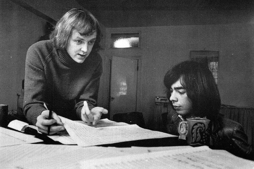 Svartvit bild av en ung Tim Rice och Andrew Lloyd Webber som arbetar med noter eller manus vid ett bord.