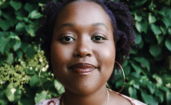 Porträttfoto på Anthia Nigitegeka, stående framför ett stort grön buske leende med blick in mot kameran. Foto taget av Tharcilla Iteka