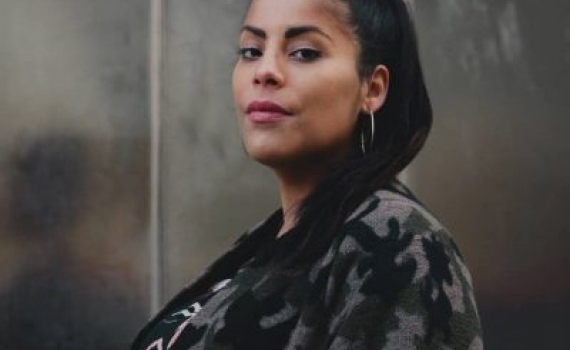 Porträttfoto av Emma Dominguez, stående vänd snett åt vänster med blicken in i kameran, mot en grå metallisk vägg som bakgrund. Foto taget av Maryam Dinar.