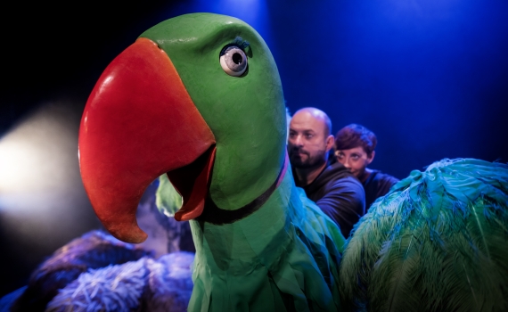 Två personer står bakom en stor papegojdocka. 