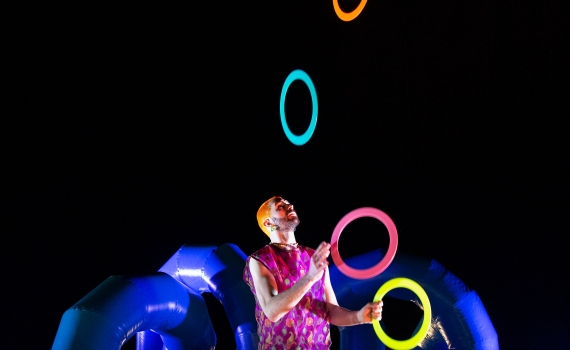 Jonglören Wes jonglerar sju plastringar i olika starka färger. Wes har rosa hår och kläder och bakom Wes syns en uppblåst plastskulptur i blått. Strålkastaljuset är på Wes 