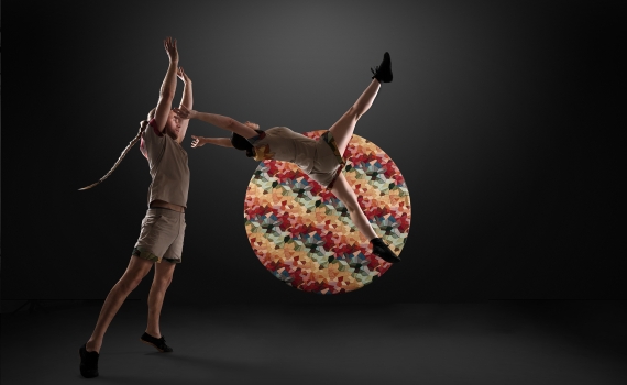 Två akrobater på en upplyst scen. Den ena kastas ur den andras armar och bakom dem är ett runt objekt med färgglatt mönster.