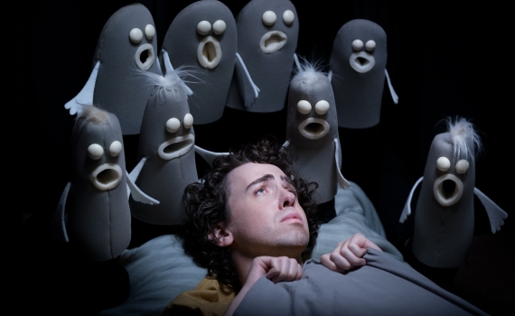 En människa ligger i en säng, omgiven av svarta och grå spökliknande dockor. 