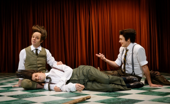 En skådespelare ligger ner på en scen, två andra sitter bredvid. 