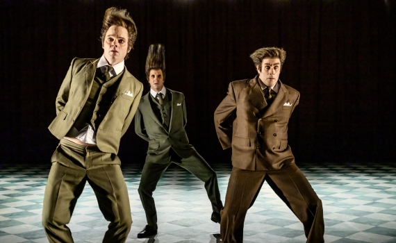 Tre skådespelare klädda i kostym ser ut att dansa på en scen. 