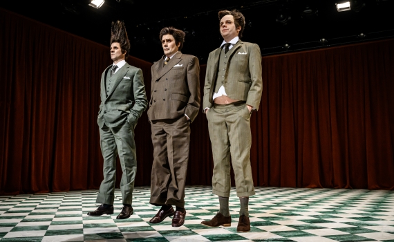 Tre skådespelare klädda i kostym hoppar på en scen. 