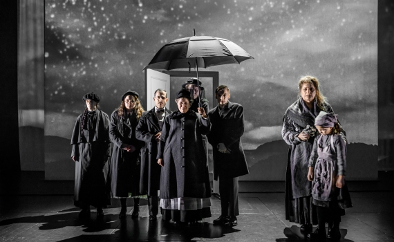 Flera skådespelare på scen i pjäsen Skuggan över stenbänken, en av dem håller i ett paraply. . 