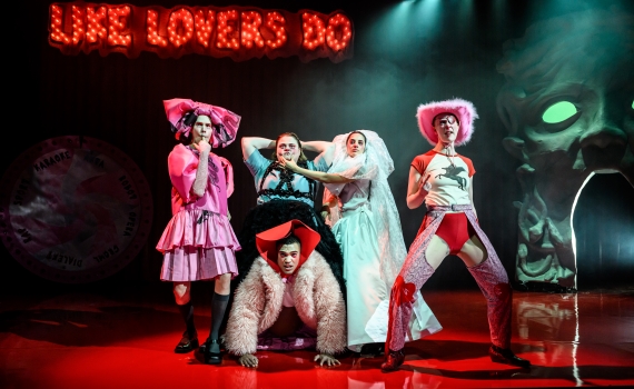 Fem skådespelare på scen klädda i fuskpälsar, rosa hattar, bröllopsklänning med mera. 