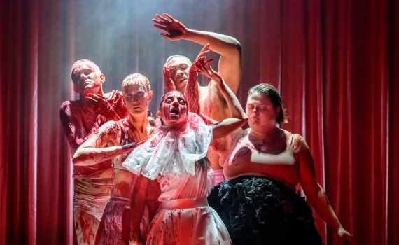 Fem skådespelare på scen täckta i blod, de blundar och sträcker sina armar uppåt. 