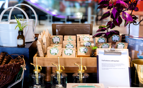 Färgglad butikshörna med fröpåsar, vattenkannor och odlingstillbehör. En lapp med odlingstips syns framträdande bland produkterna.