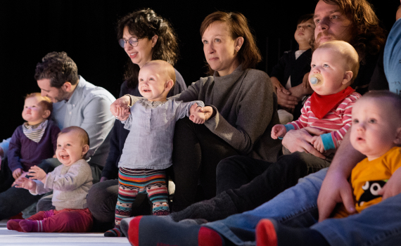 Vuxna och spädbarn sitter på golvet i publik, flera barn hålls i famn eller står med stöd.