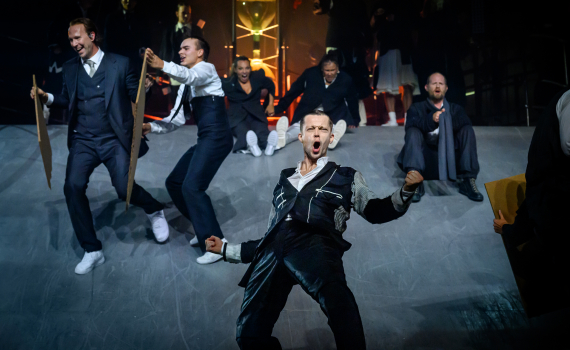 Foto: Sören Vilks Flera vuxna i scenkläder dansar på lutande scen, en man står främst och ropar med öppen mun.