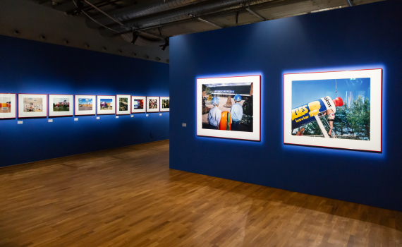 Inramade färgfoton upphängda på blå vägg i ett galleri med trägolv.