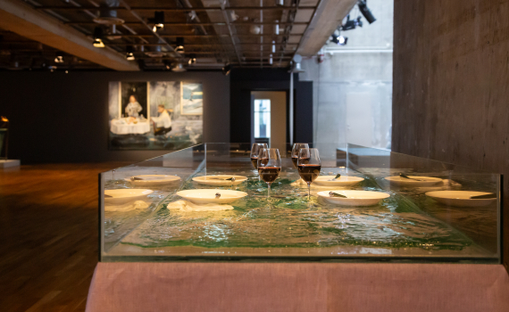 Glasbord med tallrikar, bestick och vinglas, placerade ovanpå en konstgjord vattenyta i ett galleri.