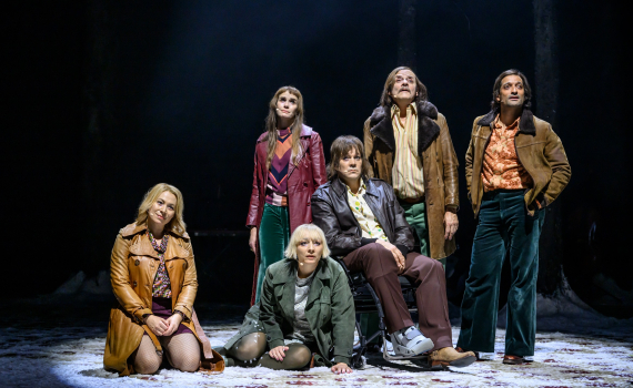 Sex vuxna i 70-talskläder poserar på scen med snö och mörk bakgrund, en man sitter i rullstol.