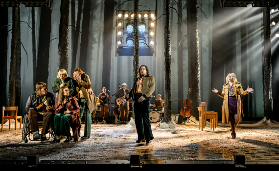 Skådespelare i 70-talskläder framför en scen i en skogsmiljö med levande band i bakgrunden.