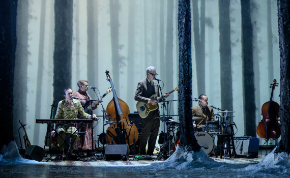 Fyra musiker spelar på scen med instrument som elgitarr, kontrabas och trummor, skog i bakgrunden.
