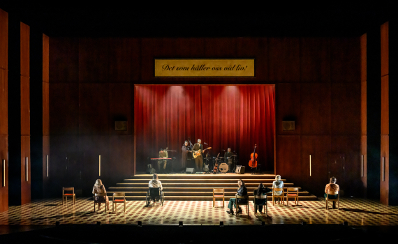 Åtta personer sitter utspridda på scen framför ett band, ovanför hänger skylten "Det som håller oss vid liv!".